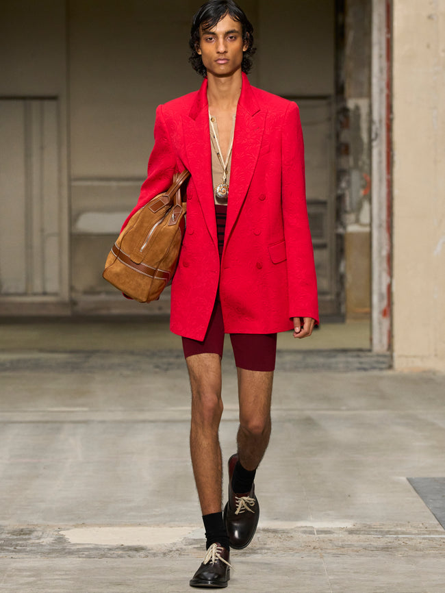 Model wearing a bright red blazer with a brown bag on a runway. - Dries Van Noten