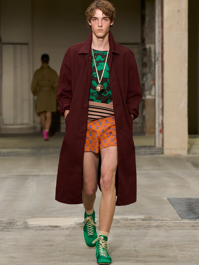 Model walking on a runway wearing a long dark coat, patterned shorts, and green shoes. - Dries Van Noten