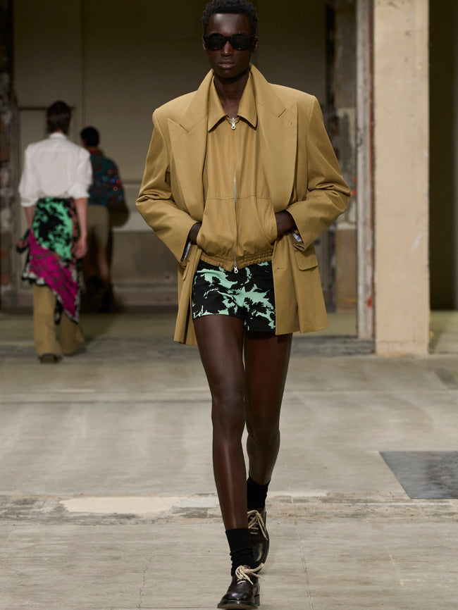 Model walking on a runway wearing a beige coat and patterned shorts. - Dries Van Noten
