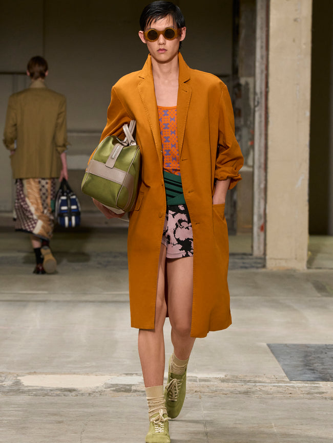 Model wearing a mustard coat and sunglasses, holding a green bag on a runway. - Dries Van Noten