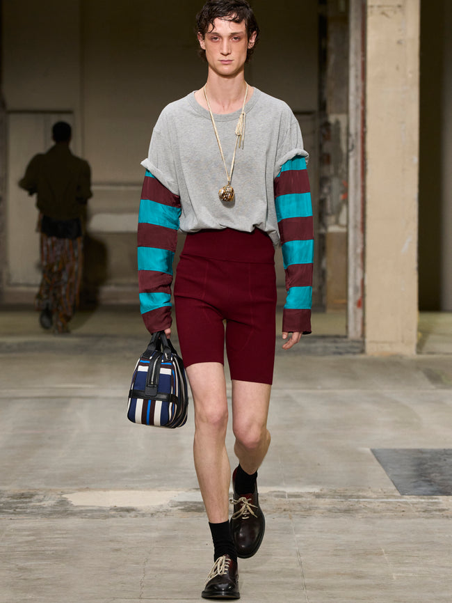 Model walking on a runway wearing a gray sweater with colorful stripes, maroon shorts, and carrying a striped bag. - Dries Van Noten