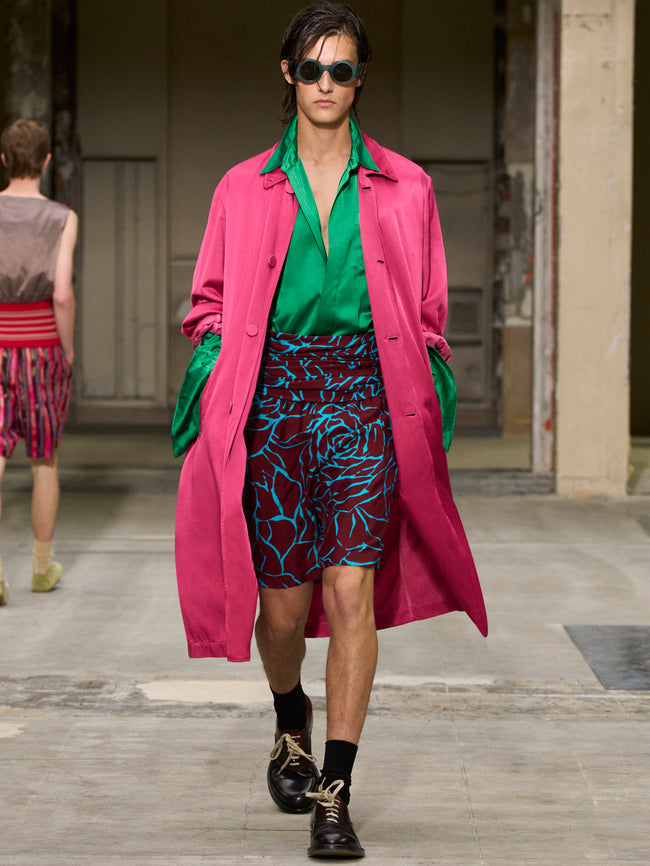 Model walking on a runway wearing a pink coat, green shirt, and patterned shorts. - Dries Van Noten