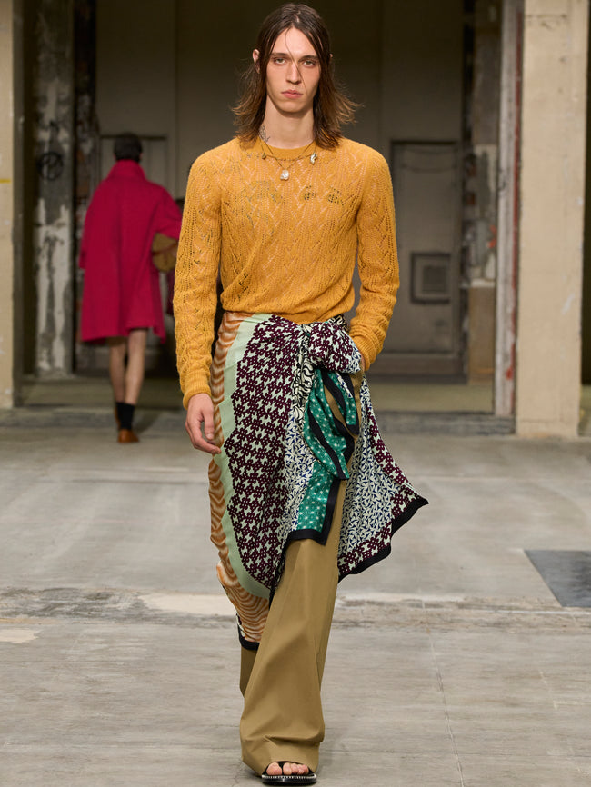 Model walking on a runway wearing a mustard yellow sweater and patterned scarf. - Dries Van Noten
