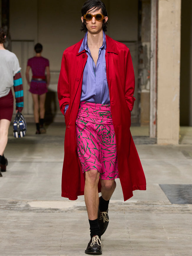 Model wearing a red coat, purple shirt, and pink shorts on a runway. - Dries Van Noten