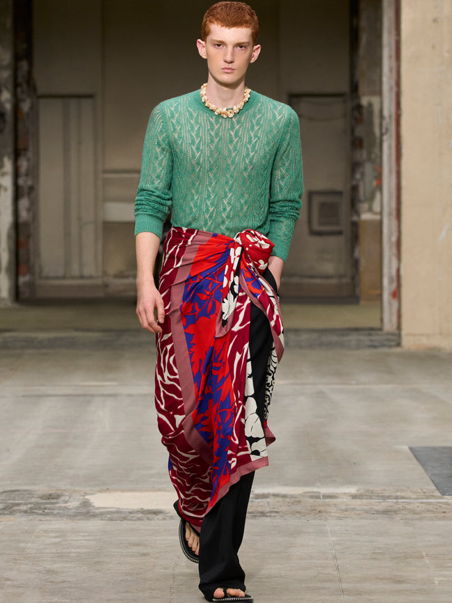 Man wearing a green sweater and red patterned skirt on a wooden floor. - Dries Van Noten