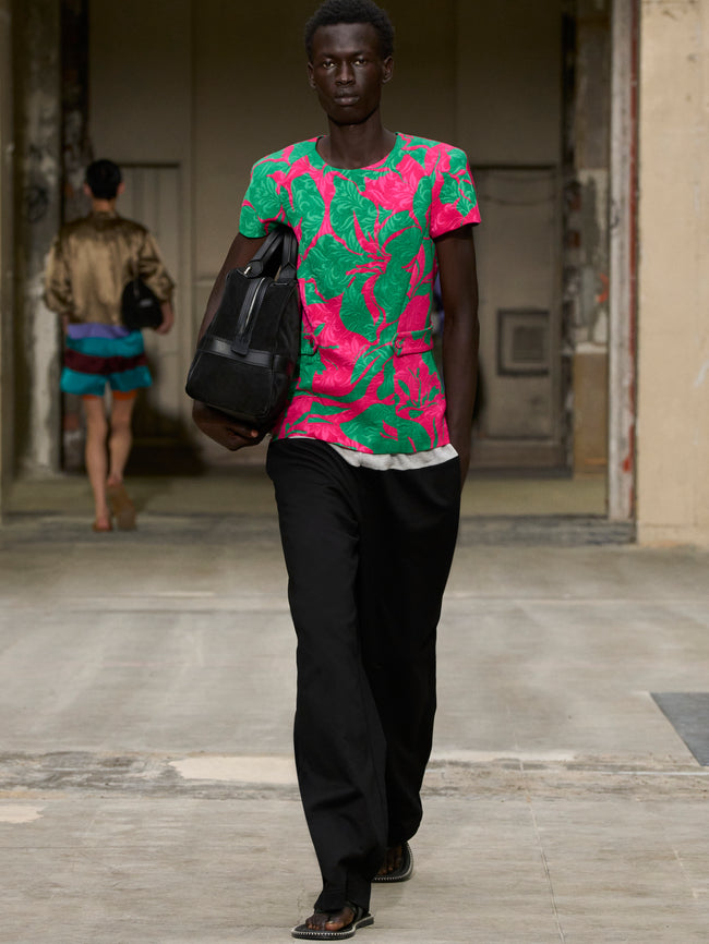 Man wearing a pink and green floral shirt with black pants, holding a black bag. - Dries Van Noten