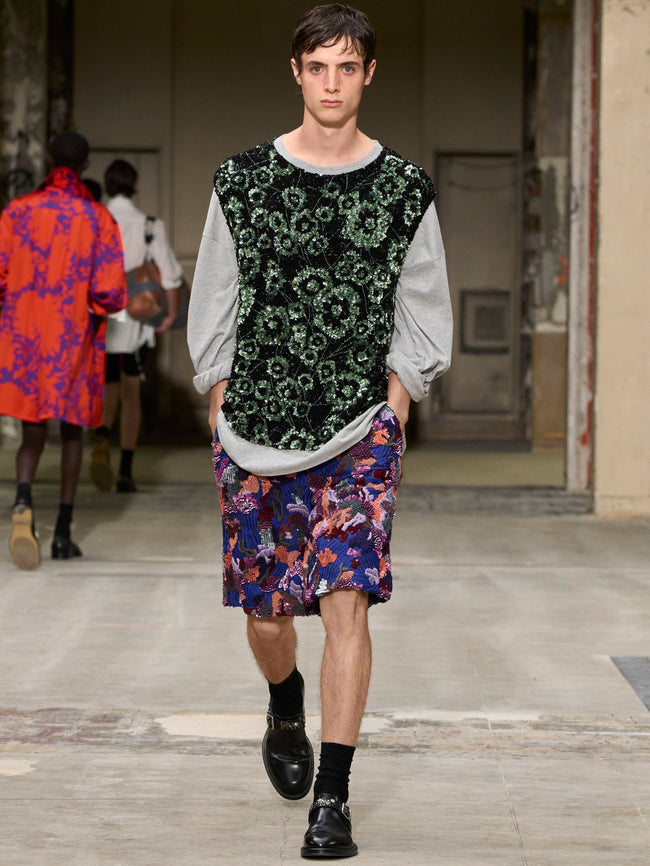 Model walking on a runway wearing a patterned outfit with a floral skirt and black top. - Dries Van Noten