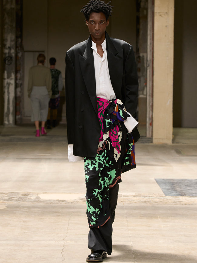 Model wearing a black coat, white shirt, and colorful pants walking outdoors. - Dries Van Noten