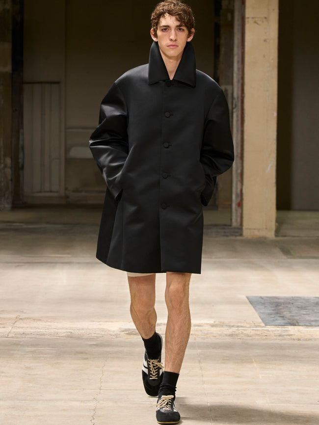 Man wearing a black coat standing in an indoor setting with wooden floors and a neutral color palette. - Dries Van Noten