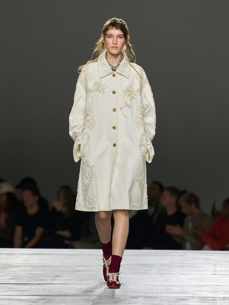 Model wearing a white embroidered coat on a runway with a dark background