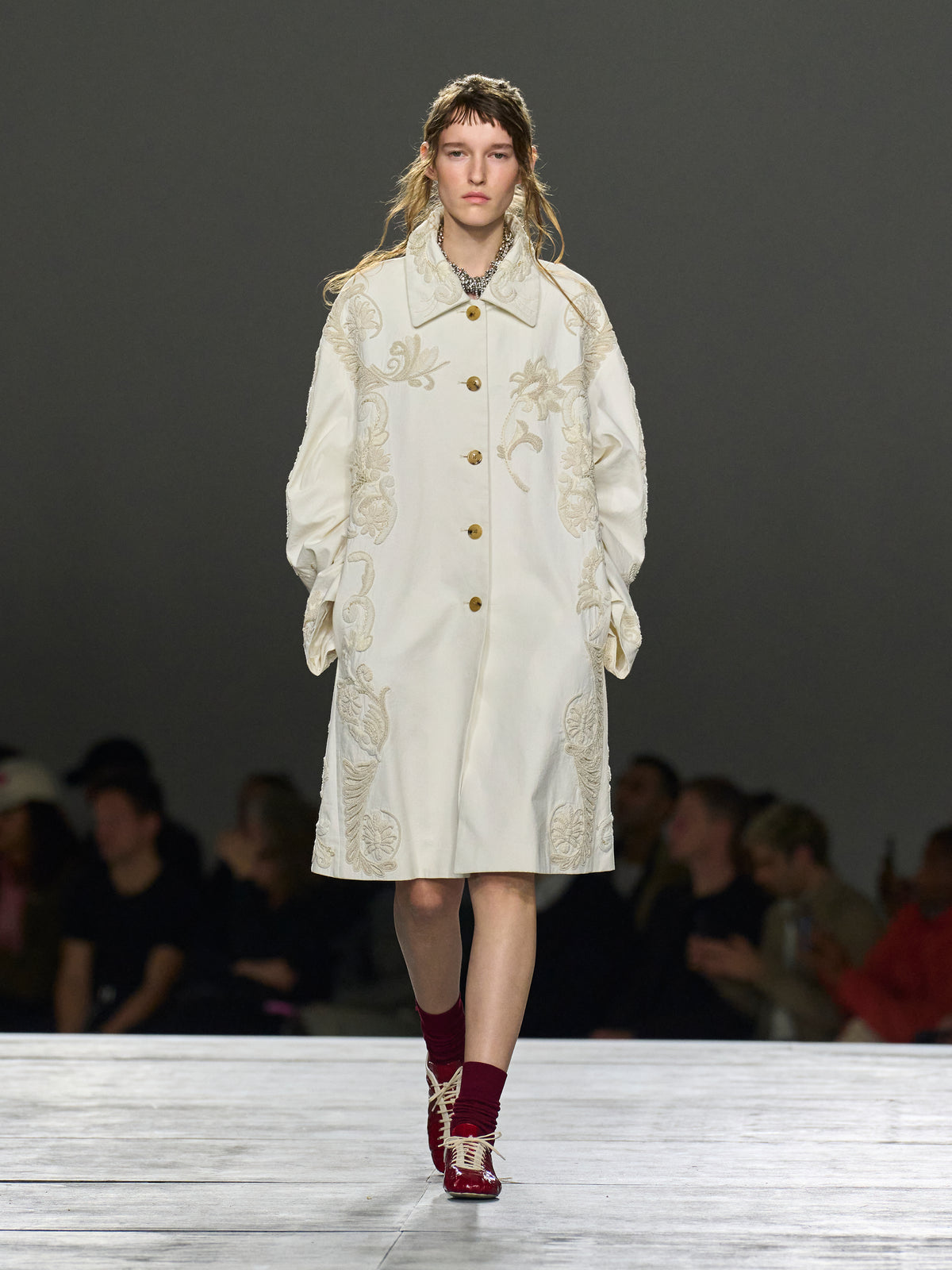 Model wearing a white embroidered coat on a runway with a dark background