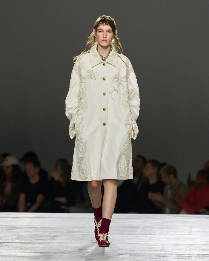 Model wearing a white embroidered coat on a runway with a dark background