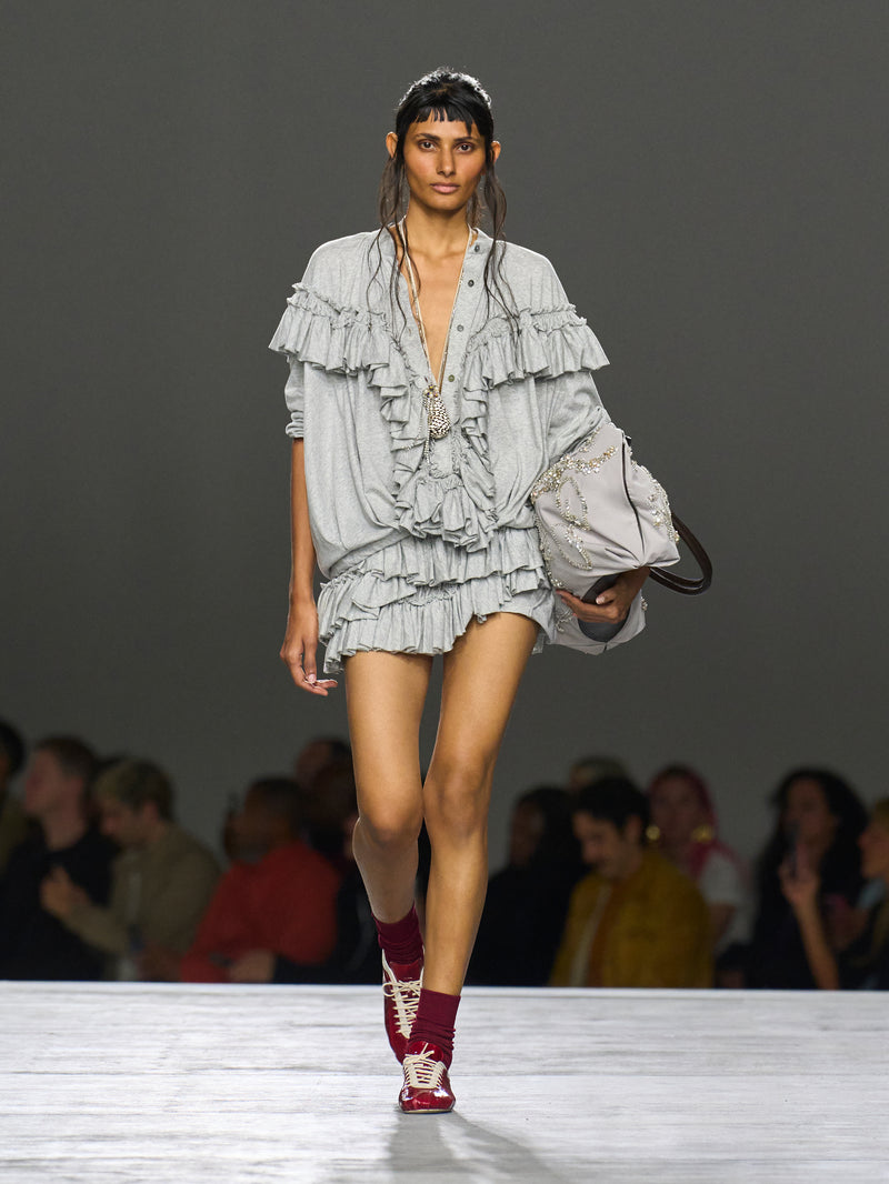 Model walking on a runway wearing a layered ruffled outfit with red shoes.