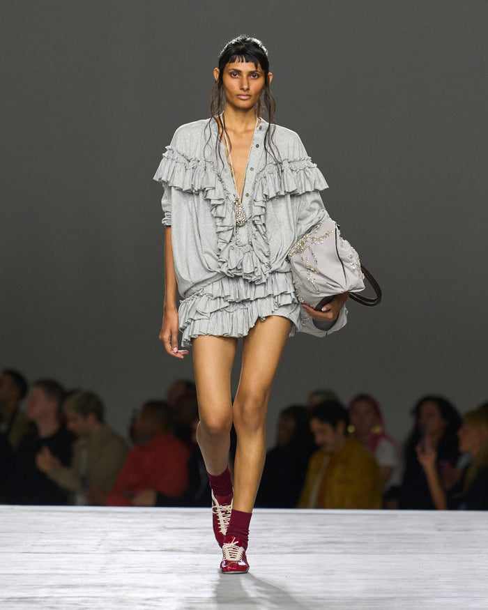 Model walking on a runway wearing a layered ruffled outfit with red shoes.