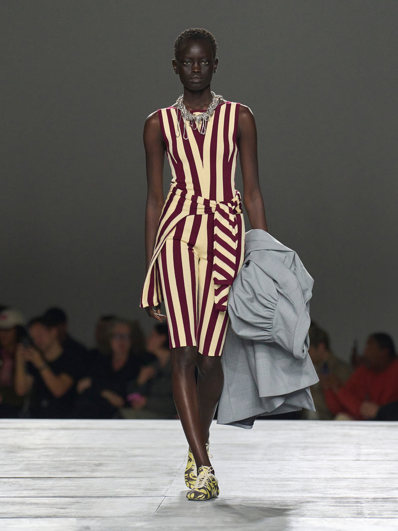 Model walking on a runway wearing a striped dress with a gray jacket.