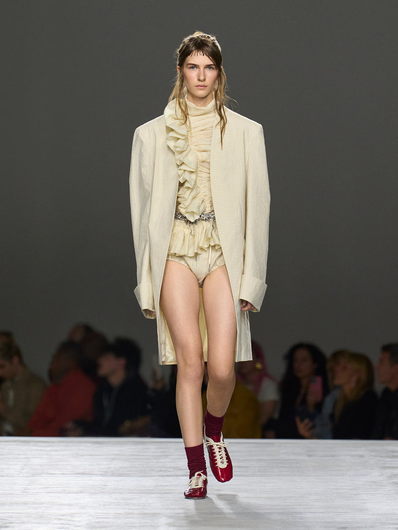 Model walking on a runway wearing a cream-colored outfit with a long coat.