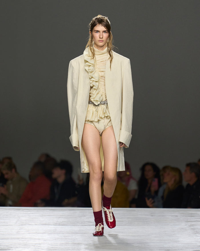Model walking on a runway wearing a cream-colored outfit with a long coat.