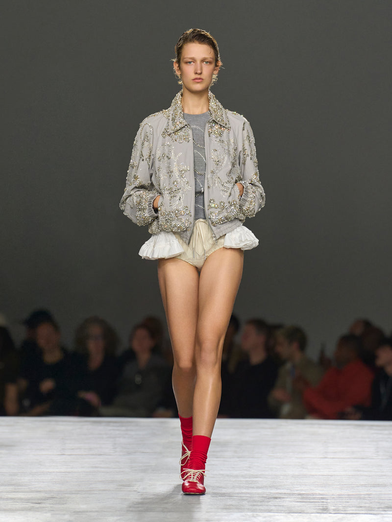 Model walking on a runway wearing a floral jacket, gray top, beige shorts, and red shoes.
