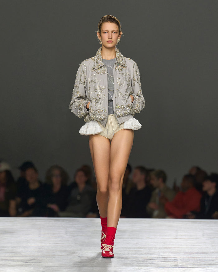 Model walking on a runway wearing a floral jacket, gray top, beige shorts, and red shoes.