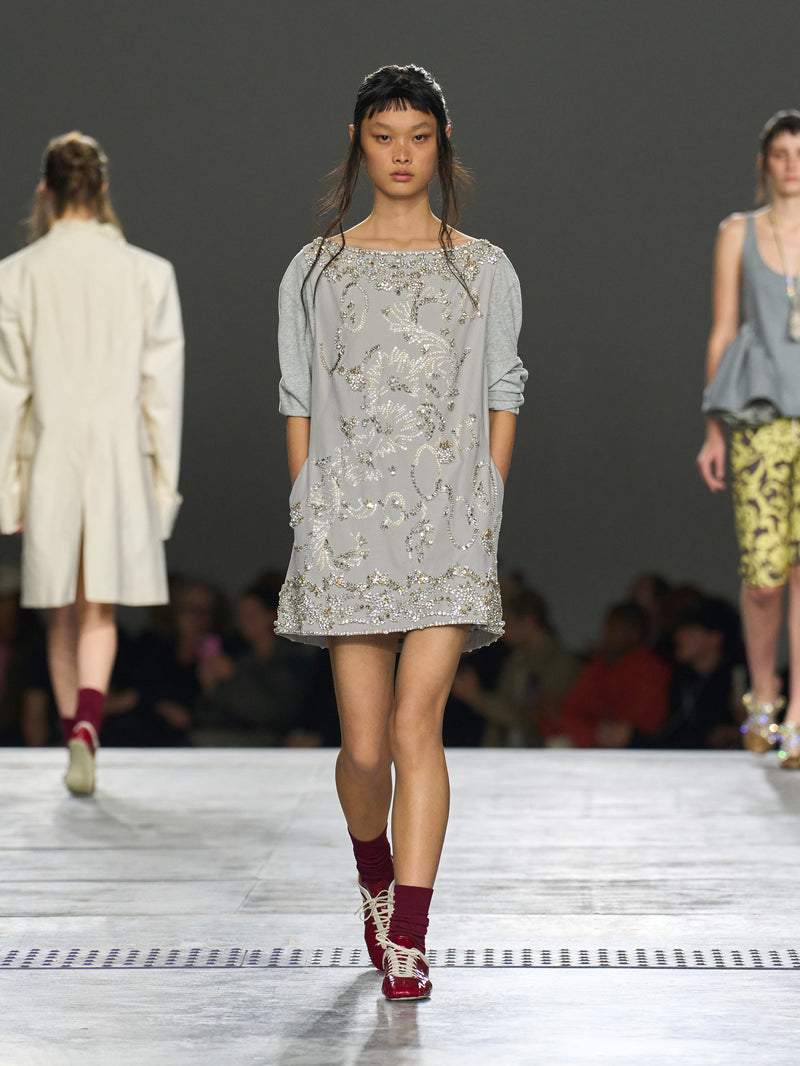 Model walking on a runway wearing a light gray dress with floral patterns.