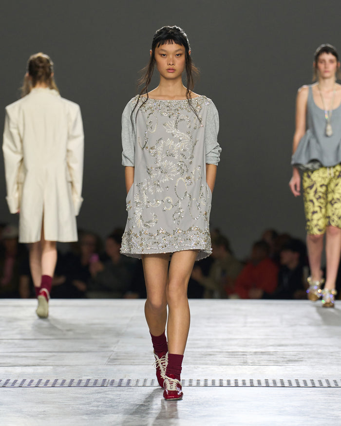 Model walking on a runway wearing a light gray dress with floral patterns.