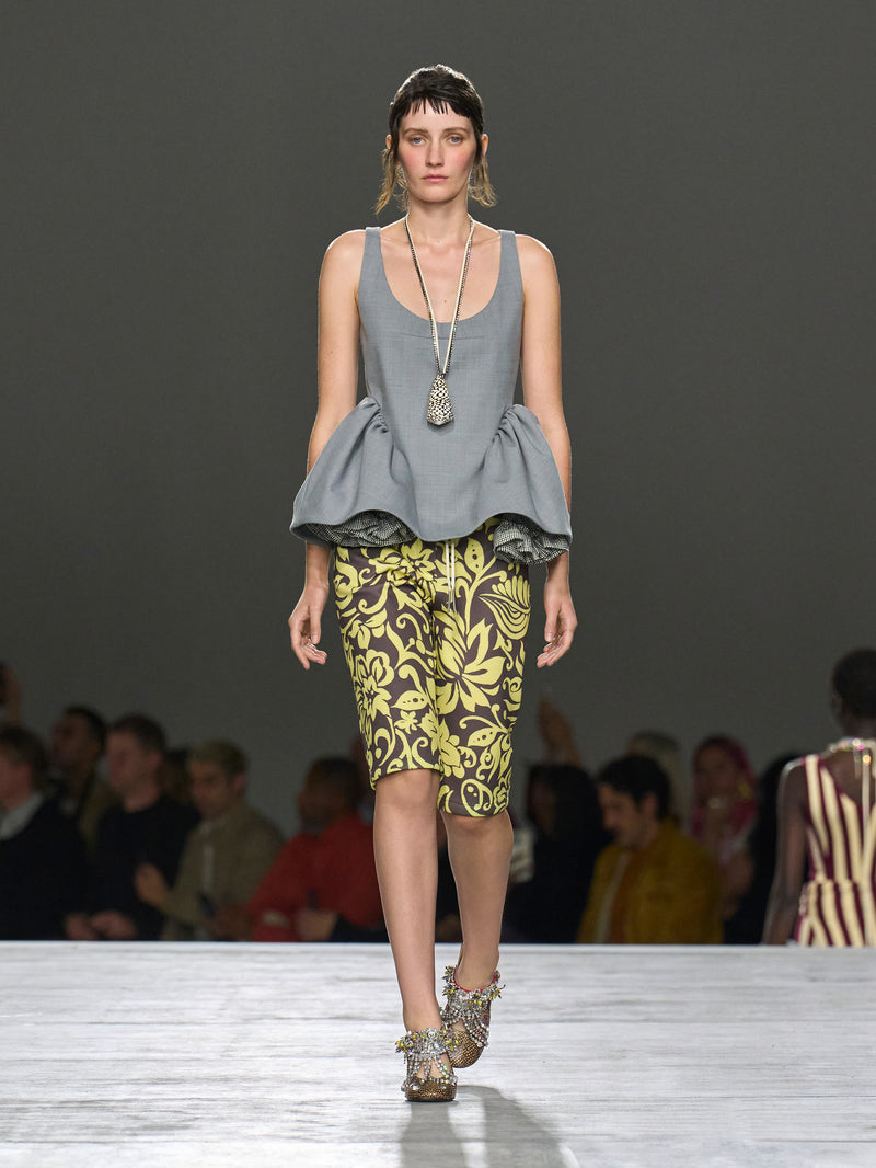 Model walking on a runway wearing a gray peplum top and patterned skirt.