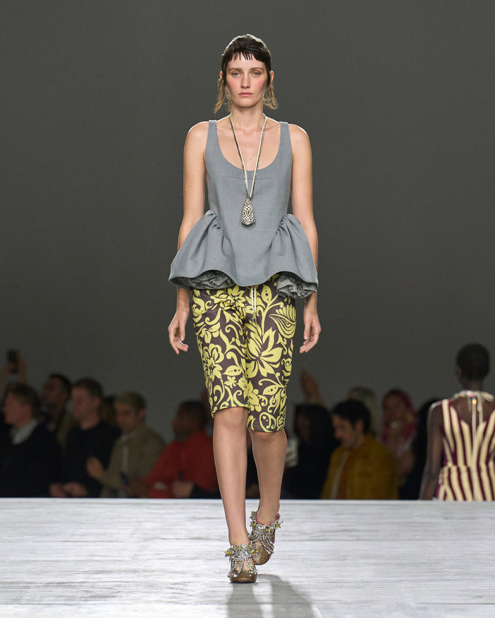 Model walking on a runway wearing a gray peplum top and patterned skirt.