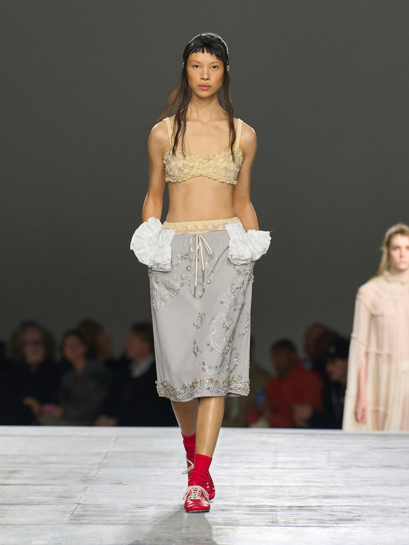 Model walking on a runway wearing a beige top and floral skirt with red shoes.