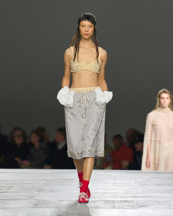 Model walking on a runway wearing a beige top and floral skirt with red shoes.