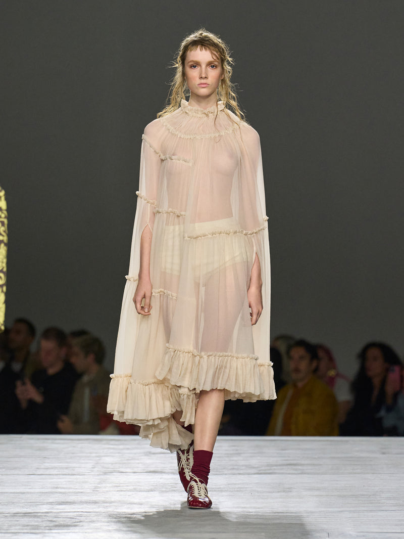 Model wearing a sheer, layered dress on a runway with an audience in the background.