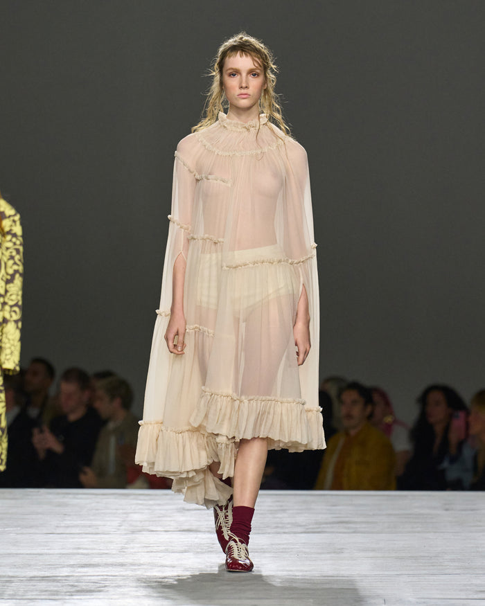 Model wearing a sheer, layered dress on a runway with an audience in the background.