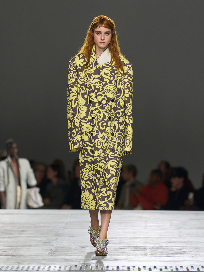 Model wearing a yellow and black patterned outfit on a runway with an audience.