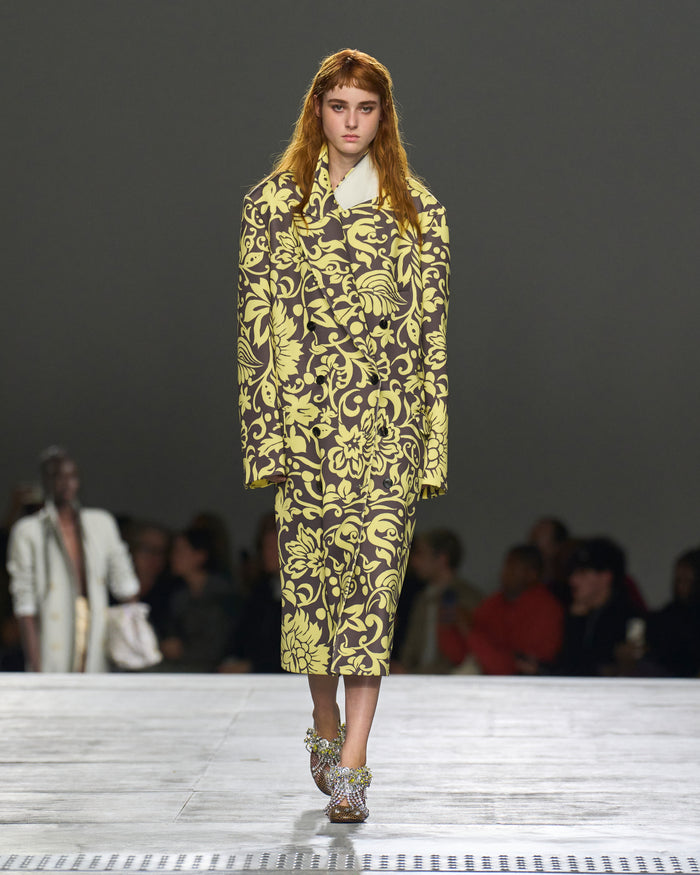 Model wearing a yellow and black patterned outfit on a runway with an audience.