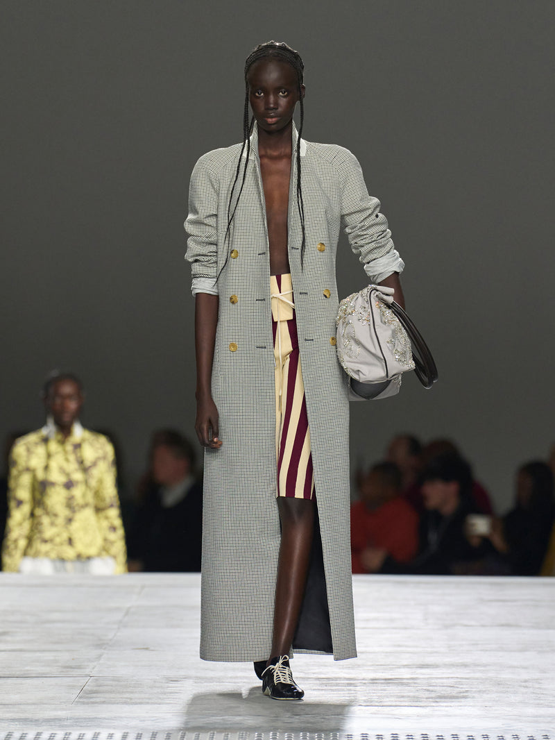 Model walking on a runway wearing a long gray coat with a patterned dress underneath.