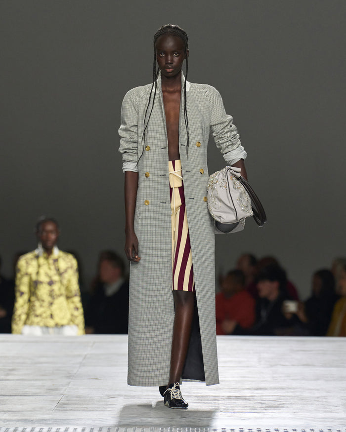Model walking on a runway wearing a long gray coat with a patterned dress underneath.