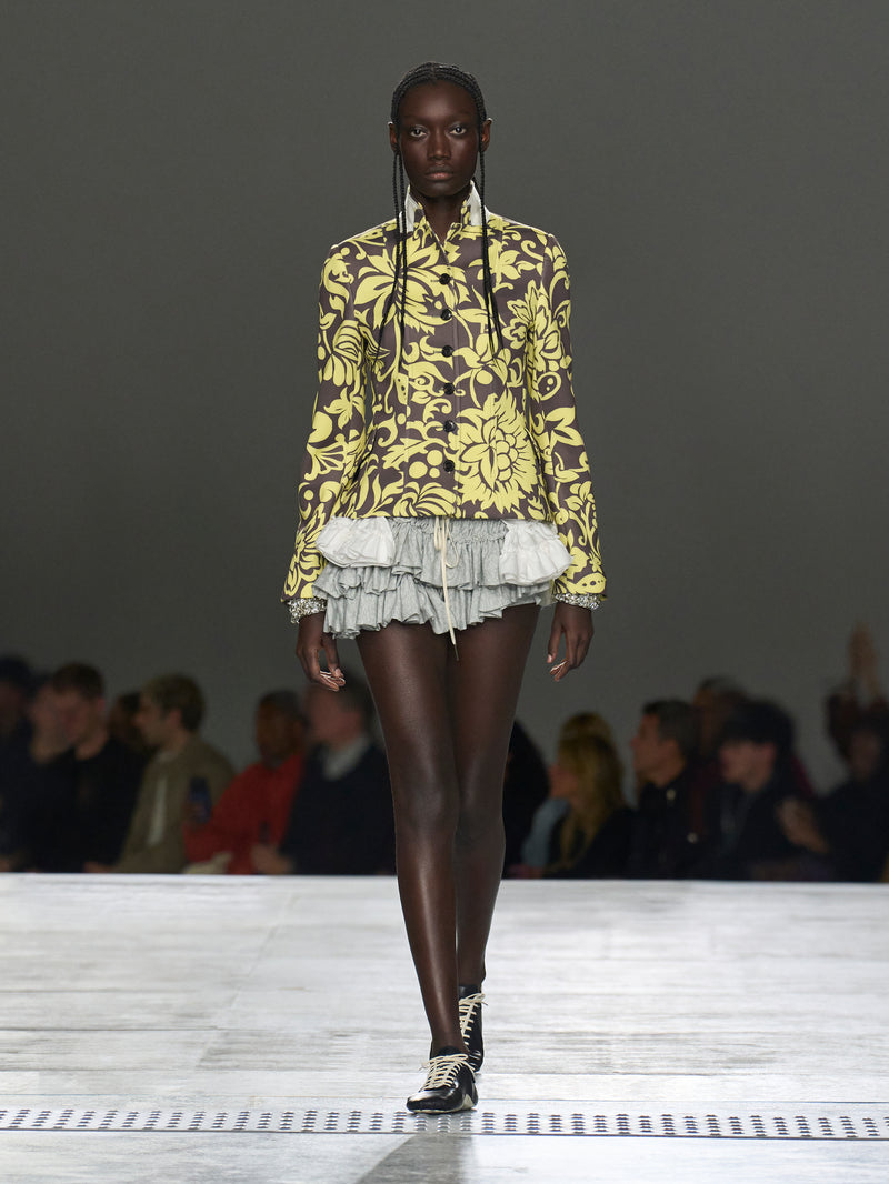 Model walking on a runway wearing a yellow floral blouse and gray pleated skirt.