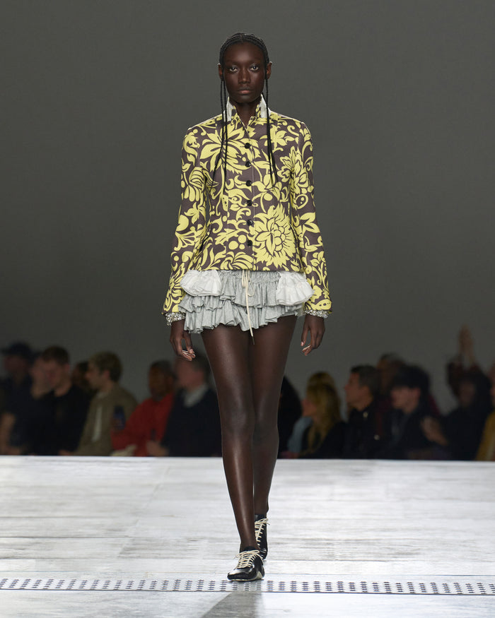 Model walking on a runway wearing a yellow floral blouse and gray pleated skirt.
