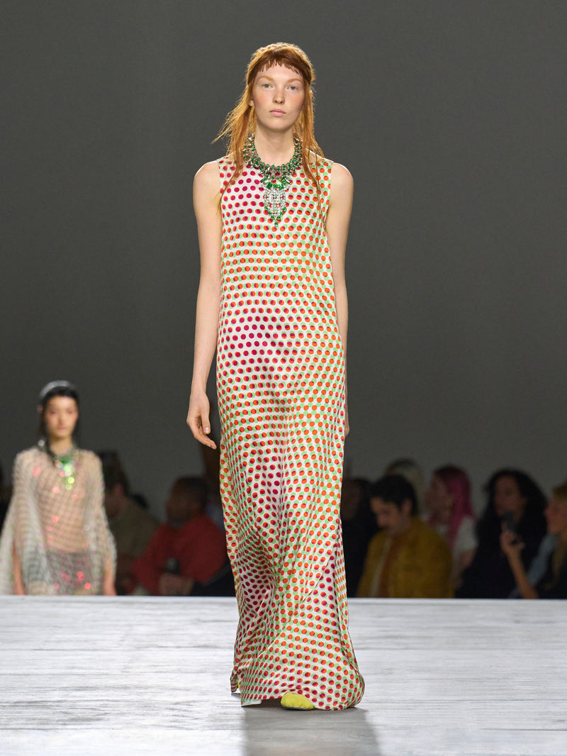 Model walking on a runway wearing a patterned dress with a dark background