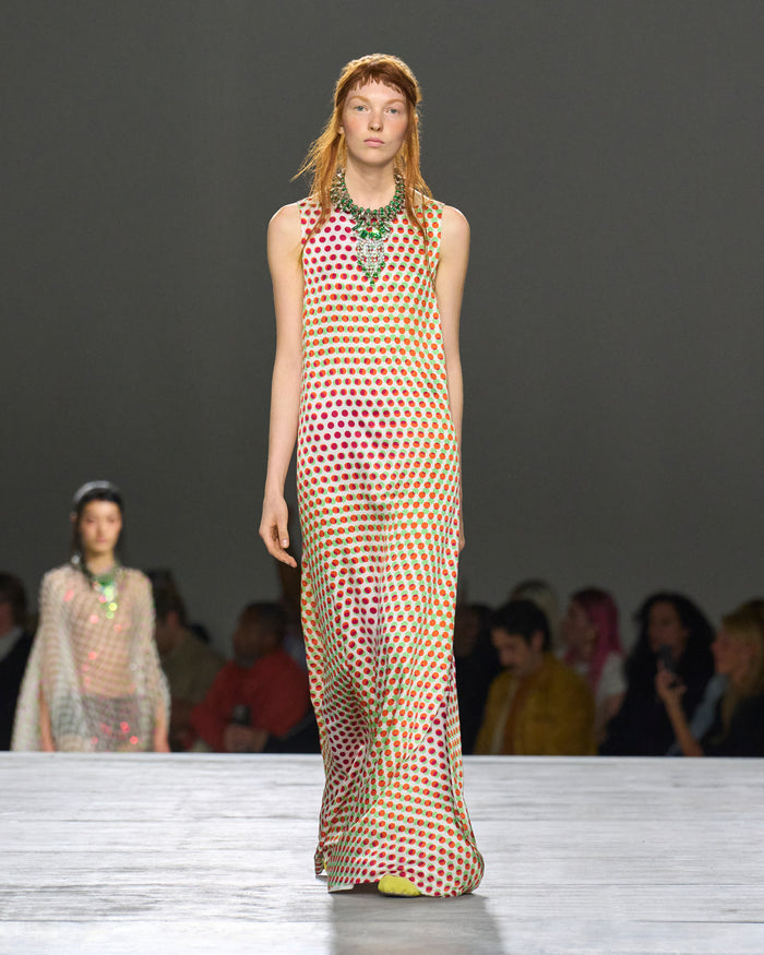 Model walking on a runway wearing a patterned dress with a dark background