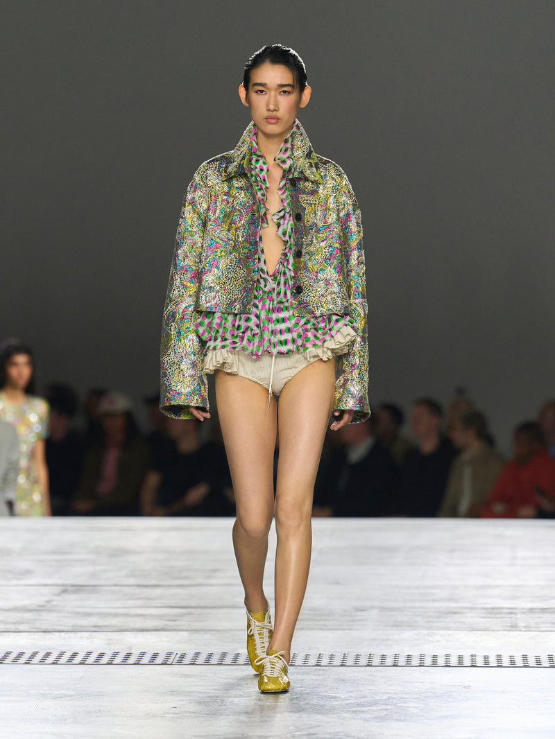 Model walking on a runway wearing a patterned outfit with a blurred audience in the background