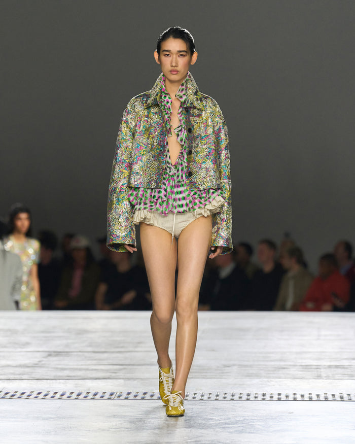 Model walking on a runway wearing a patterned outfit with a blurred audience in the background