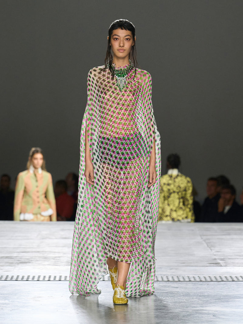 Model walking on a runway wearing a patterned dress with a dark background