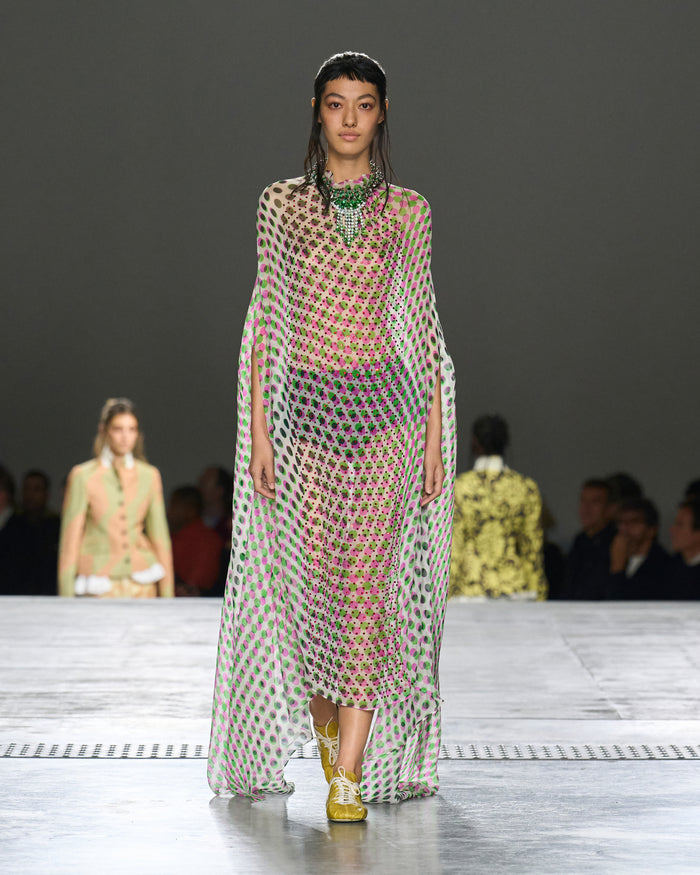 Model walking on a runway wearing a patterned dress with a dark background