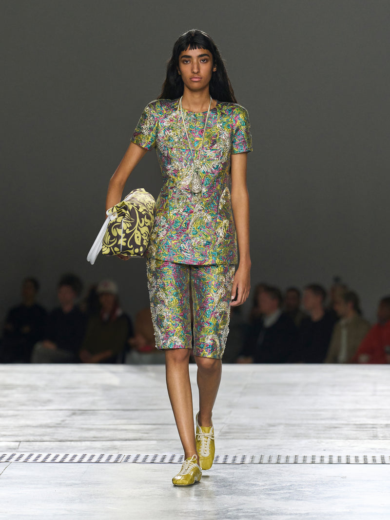 Model walking on a runway wearing a colorful outfit with a handbag.