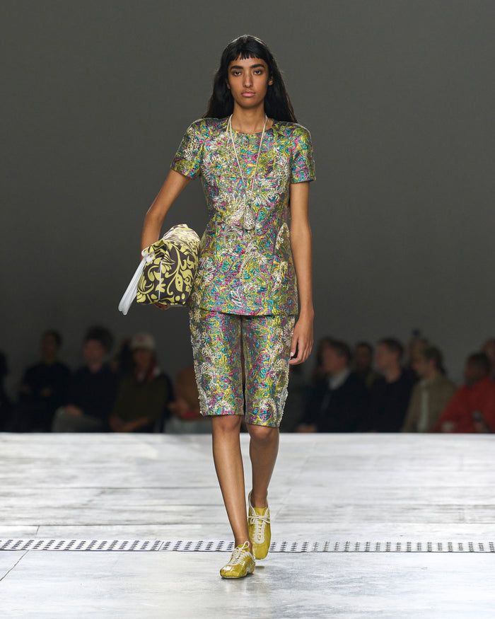 Model walking on a runway wearing a colorful outfit with a handbag.