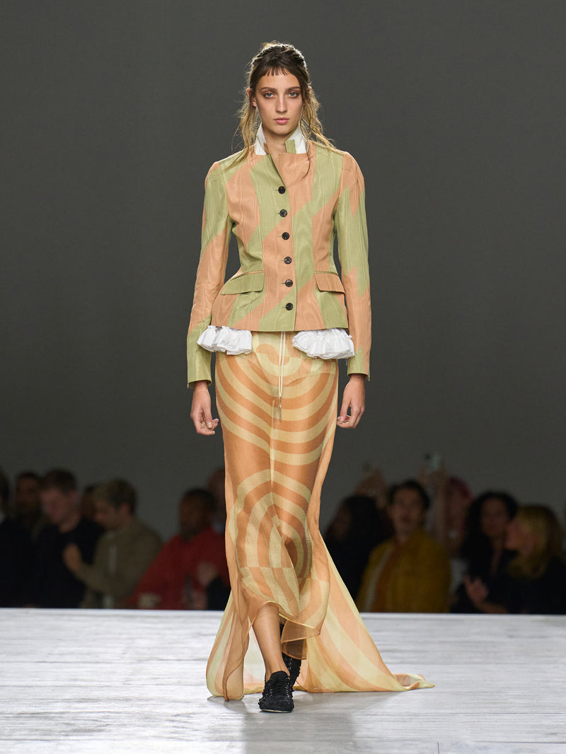 Model wearing a patterned outfit on a runway with an audience in the background.