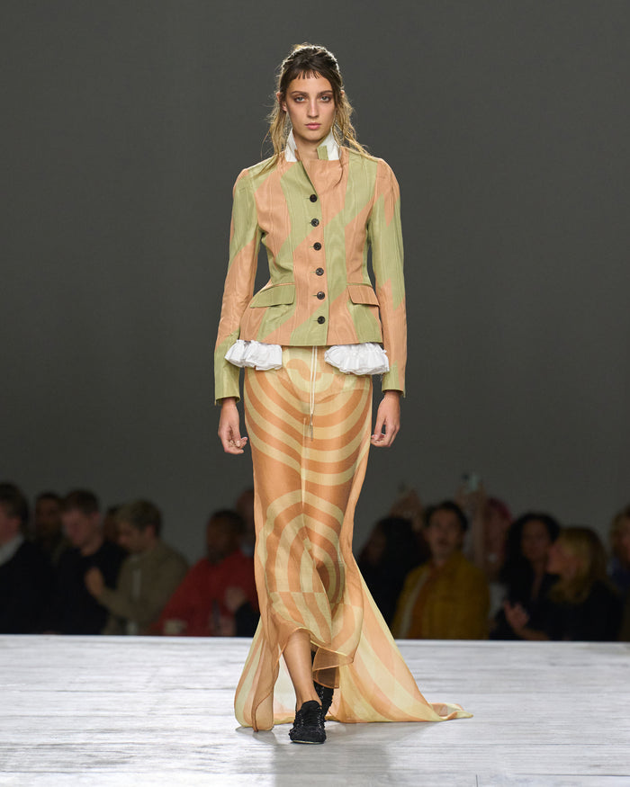 Model wearing a patterned outfit on a runway with an audience in the background.