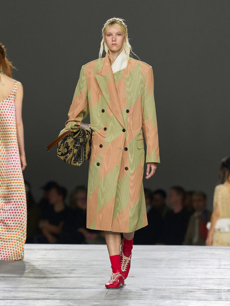 Model walking on a runway wearing a patterned coat and red shoes.