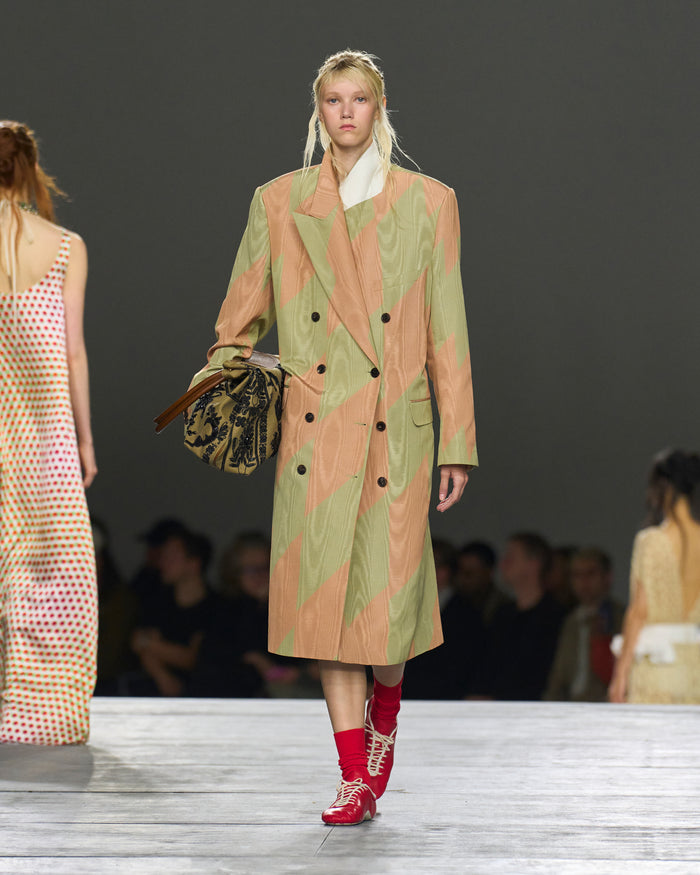 Model walking on a runway wearing a patterned coat and red shoes.