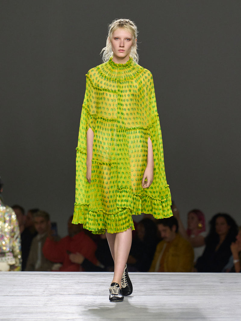 Model wearing a bright green dress on a runway with a blurred audience in the background.